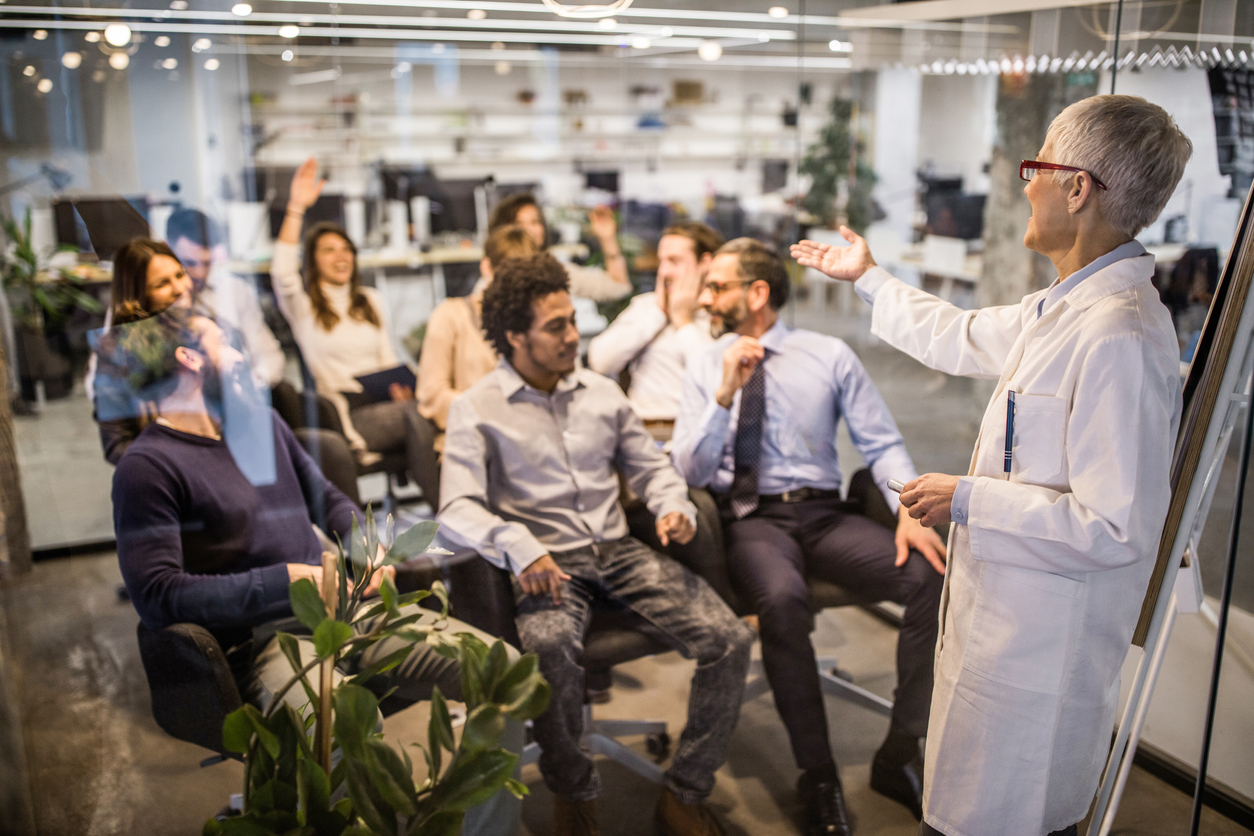 Mature female doctor teaching a seminar to large group of business people in a board room and pointing at businesswoman with raised hand. The view is through the glass.